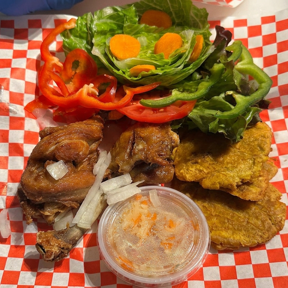 Jamaican-style platter with jerk chicken, fried plantains, fresh salad, sliced peppers, onions, and dipping sauce on checkered paper.